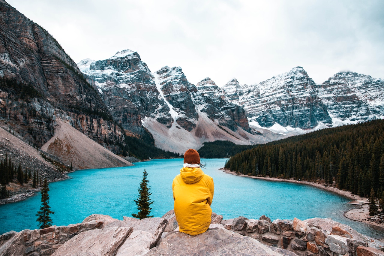 person in yellow hoodie sitting on rock near lake and snow covered mountain during daytime